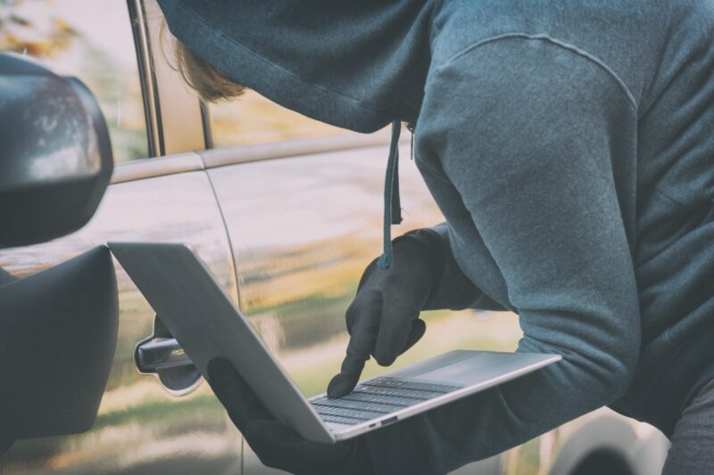 Un uomo con cappuccio e guanti neri manovra un computer portatile accanto alla maniglia di un'auto.